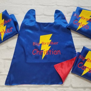 May include: Blue satin superhero capes and matching masks with yellow lightning bolt designs. The capes and masks have the words "Big Brother" and individual names in red. The cape is reversible with a red interior.