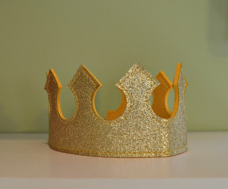 May include: A gold glitter crown with a yellow interior. The crown has a classic design with pointed arches and a rounded base. The glitter adds a festive touch, making it suitable for celebrations or dress-up.