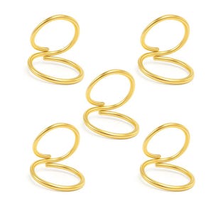 May include: Five gold-colored, double-loop rings arranged on a white background. Each ring has a unique, open design with two connected circular loops. The rings are made of a shiny, metallic material.
