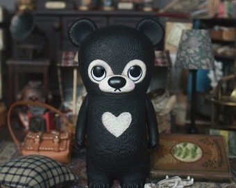 May include: A black bear figurine with a white heart on its chest. The bear has large, round eyes and a small, black nose. The figurine is standing on a patterned rug in a miniature room setting.