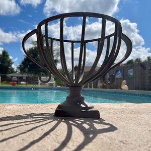 May include: A metal planter in the shape of a trophy cup with a decorative design. The planter is sitting on a concrete surface near a swimming pool.