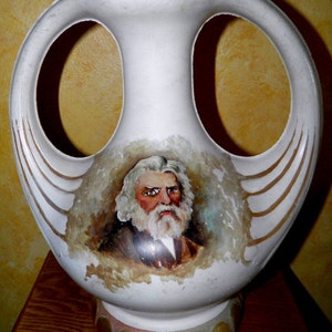 May include: White ceramic vase with two handles and a painted portrait of a man with a beard. The vase has a gold and green trim and a base with gold dots.