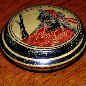 May include: A round, black and gold trinket box with a red and black painted design of a figure with the inscription "ZEYS KALOS".