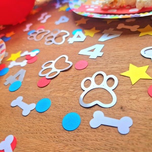 May include: A wooden table is scattered with party decorations, including silver paw prints, white bone shapes, yellow stars, red and blue circles, and the number 4. A red balloon and a colorful plate are also visible.