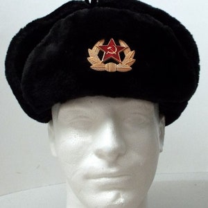 Puede incluir: Un gorro ushanka de piel sintética negra con una estrella roja y un emblema de martillo y hoz.