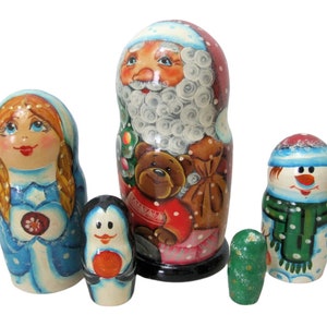 Pode incluir: Um conjunto de cinco bonecas russas tradicionais, ou bonecas matrioska, decoradas com um tema de Natal de inverno. A boneca maior representa o Papai Noel com barba branca, segurando um urso de pelúcia. A segunda boneca maior é uma mulher com cabelo longo e loiro que usa um vestido azul. A terceira boneca maior é um pinguim que segura uma bola vermelha. A quarta boneca maior é um boneco de neve que usa uma echarpe e um chapéu verdes. A boneca menor é verde.