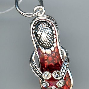 May include: Silver flip-flop charm with a red enamel base and clear crystal accents. The charm has a textured sole and a loop for attaching to a necklace or bracelet. A summer-themed jewelry item.