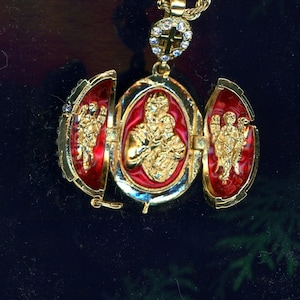 May include: A gold pendant necklace with a red and gold egg-shaped charm. The charm is open to reveal three smaller compartments, each with a gold and red image of a religious figure.