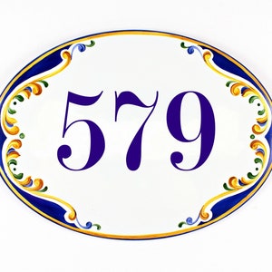 Oval ceramic house number sign with the number 579 painted in a deep purple font. The sign is hand-painted with a blue and gold decorative border. The border features ornate floral designs.