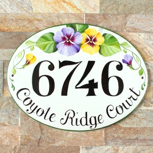 oval ceramic house number sign with a green border. The number 6746 is painted in black. The sign is hand painted with purple and yellow pansies with green leaves. The text "Coyote Ridge Court" is written in black cursive.