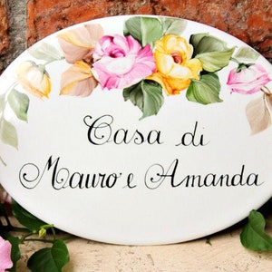 May include: White oval ceramic sign with a floral design featuring pink and yellow roses. The sign reads "Casa di Mauro e Amanda".