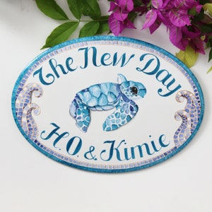 May include: Oval ceramic sign with a blue and white mosaic border. The sign features a watercolor painting of a sea turtle swimming, with the text "The New Day H.O. & Kimie" in cursive.