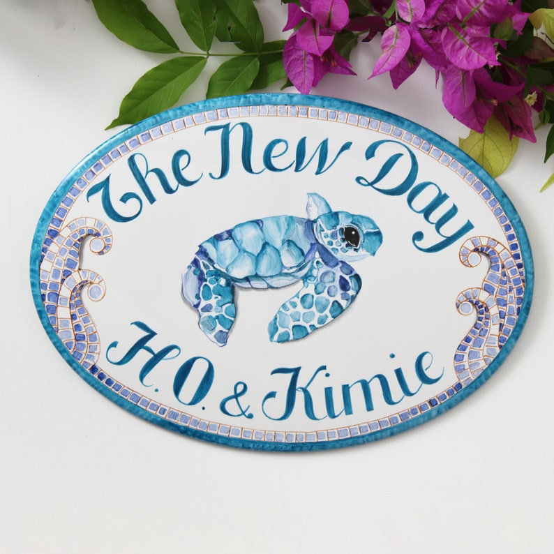 May include: Oval ceramic sign with a blue and white mosaic border. The sign features a watercolor painting of a sea turtle swimming, with the text "The New Day H.O. & Kimie" in cursive.