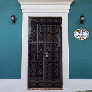 May include: A teal blue building with a white trim and a black metal door with intricate designs. The door has a white oval sign with the text "Villa Shahla" in black lettering and colorful fruit illustrations.