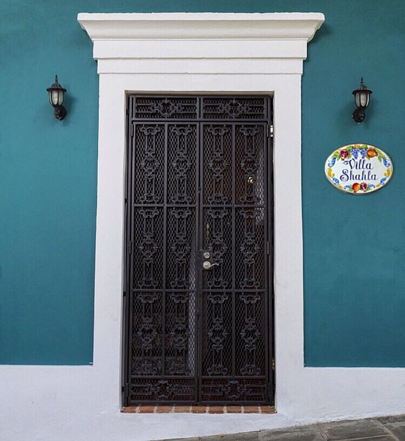May include: A teal blue building with a white trim and a black metal door with intricate designs. The door has a white oval sign with the text "Villa Shahla" in black lettering and colorful fruit illustrations.