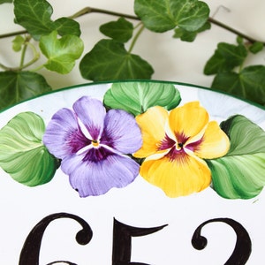white ceramic oval sign with a green border, featuring two painted purple and yellow pansies with green leaves. The number 652 is painted in black on the sign.