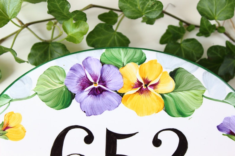 white ceramic oval sign with a green border, featuring two painted purple and yellow pansies with green leaves. The number 652 is painted in black on the sign.
