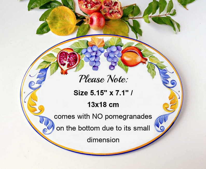 May include: Oval ceramic tile with a painted design of grapes, pomegranates, and leaves. The tile is white with blue and yellow accents. The text "Please Note: Size 5.15" x 7.1" / 13x18 cm comes with NO pomegranades on the bottom due to its small dimension" is printed on the tile.