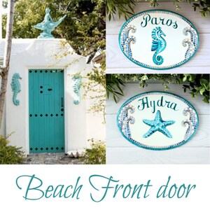May include: A turquoise blue front door with a white frame and two seahorse door knockers. Two oval-shaped mosaic signs with seahorse and starfish designs are displayed above the door. The signs read "Paros" and "Hydra".