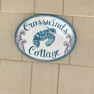 May include: White oval sign with blue and gold mosaic trim. The sign says "Crosswinds Cottage" in cursive script with a blue sea turtle illustration.