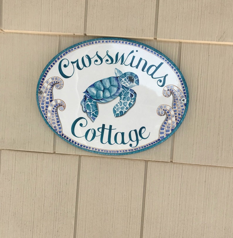 May include: White oval sign with blue and gold mosaic trim. The sign says "Crosswinds Cottage" in cursive script with a blue sea turtle illustration.