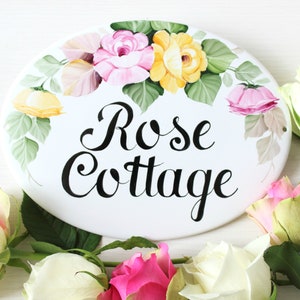 May include: White oval ceramic sign with a hand-painted floral design featuring pink, yellow, and green roses. The sign reads "Rose Cottage" in black cursive lettering.