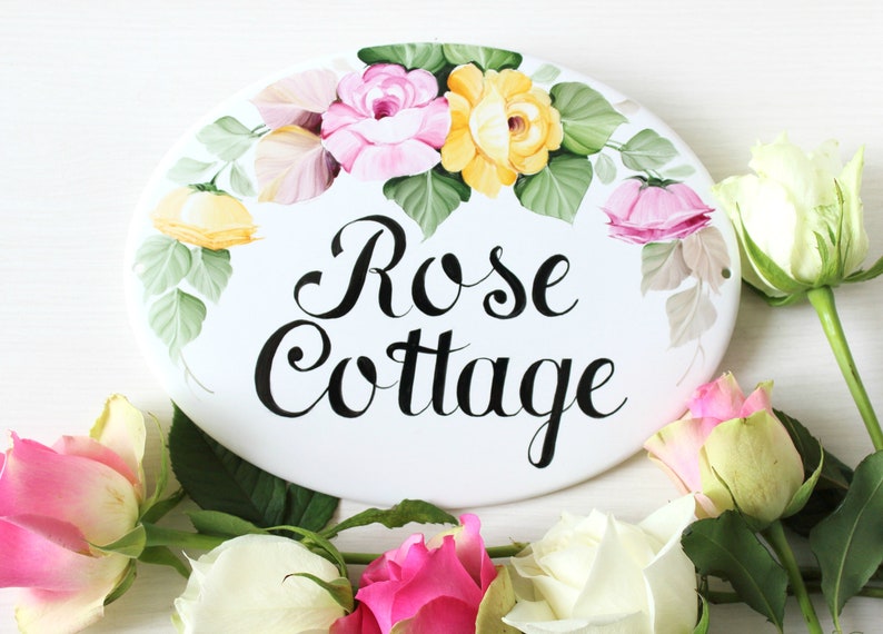 May include: White oval ceramic sign with a hand-painted floral design featuring pink, yellow, and green roses. The sign reads "Rose Cottage" in black cursive lettering.