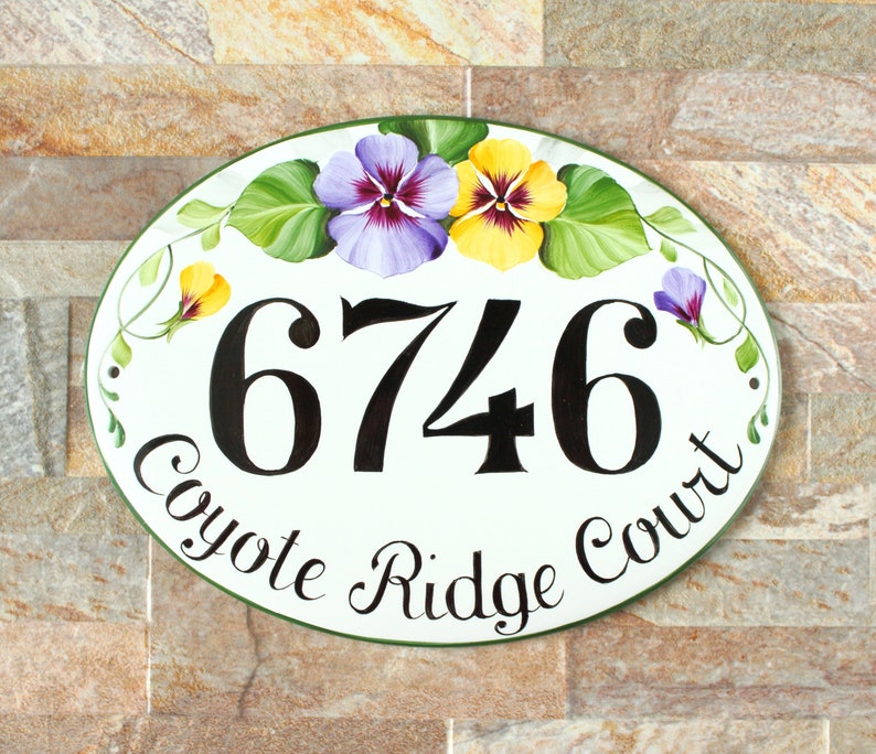 oval ceramic house number sign with a green border. The number 6746 is painted in black. The sign is hand painted with purple and yellow pansies with green leaves. The text "Coyote Ridge Court" is written in black cursive.