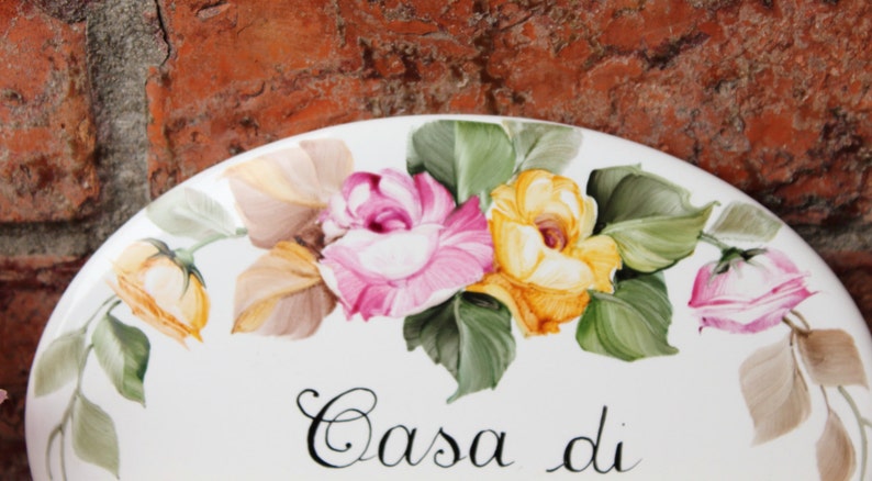 May include: White ceramic oval plate with a painted floral design of pink and yellow roses with green leaves. The plate has the text "Casa di" written in black cursive.