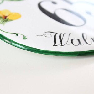 White oval ceramic house number sign with a green border. The number 65 is painted in black and the word "Walnut" is written in cursive below the number.