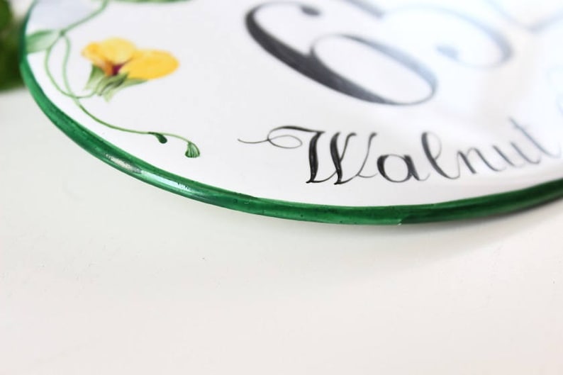 White oval ceramic house number sign with a green border. The number 65 is painted in black and the word "Walnut" is written in cursive below the number.