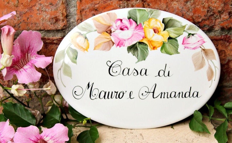 May include: White oval ceramic sign with a floral design featuring pink and yellow roses. The sign reads "Casa di Mauro e Amanda".