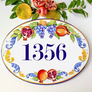 May include: Oval ceramic house number sign with a white background and blue numbers. The sign is decorated with a yellow, blue, and green floral design with red and orange pomegranates and grapes.