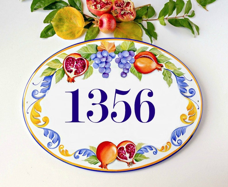 May include: Oval ceramic house number sign with a white background and blue numbers. The sign is decorated with a yellow, blue, and green floral design with red and orange pomegranates and grapes.
