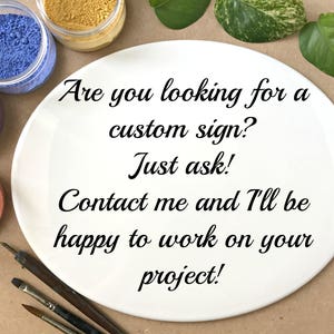 May include: A white plate with the text "Are you looking for a custom sign? Just ask! Contact me and I'll be happy to work on your project!" written in black cursive font. The plate is surrounded by small jars of colorful powder and paintbrushes.