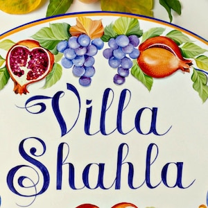May include: A white oval ceramic sign with a blue and yellow border. The sign has a painted design of grapes, pomegranates, and leaves. The text "Villa Shahla" is written in blue cursive lettering.
