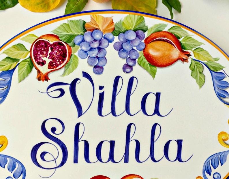 May include: A white oval ceramic sign with a blue and yellow border. The sign has a painted design of grapes, pomegranates, and leaves. The text "Villa Shahla" is written in blue cursive lettering.