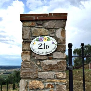 oval house number sign with a hand painted floral design and the number 21D. The sign reads "Gianluca Saccinelli Beatrice Tagliaferri". The sign is mounted on a stone wall.