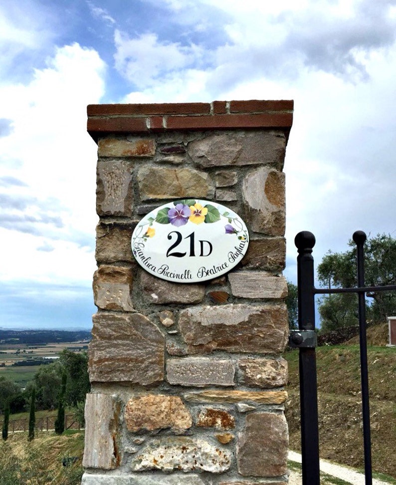 oval house number sign with a hand painted floral design and the number 21D. The sign reads "Gianluca Saccinelli Beatrice Tagliaferri". The sign is mounted on a stone wall.
