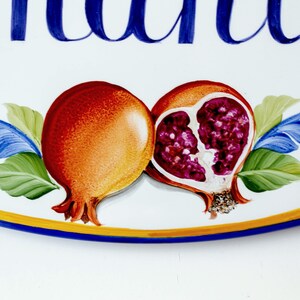May include: A white oval ceramic sign with blue lettering that reads "Giuliana". The sign is decorated with two hand-painted pomegranates, one whole and one cut open, with red and purple seeds. The pomegranates are surrounded by green leaves and blue floral accents.