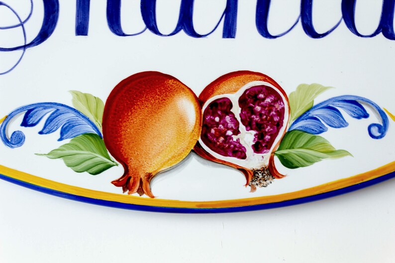 May include: A white oval ceramic sign with blue lettering that reads "Giuliana". The sign is decorated with two hand-painted pomegranates, one whole and one cut open, with red and purple seeds. The pomegranates are surrounded by green leaves and blue floral accents.