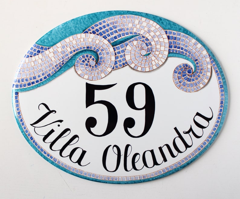 A white oval ceramic sign with a blue and white mosaic design. The sign has the number 59 and the text "Villa Oleandra" written in black cursive.