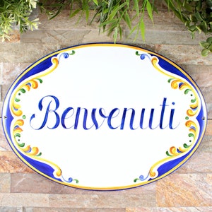 Oval ceramic sign with the word "Benvenuti" in blue script. The sign has a white background with a blue border and colorful floral accents in yellow, green, and orange. Decorative wall art.