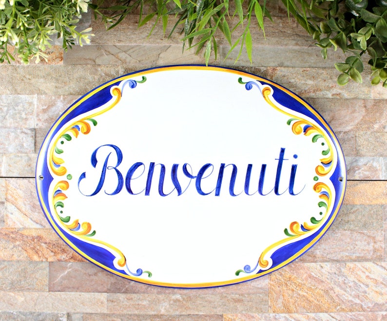 Oval ceramic sign with the word "Benvenuti" in blue script. The sign has a white background with a blue border and colorful floral accents in yellow, green, and orange. Decorative wall art.