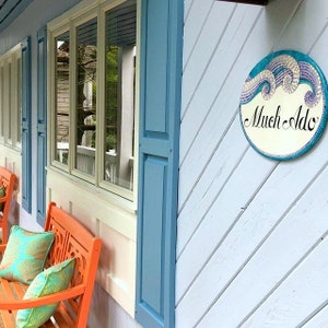 May include: A blue house with white trim and a blue shutter. There are two orange benches with cushions on the porch. A round sign with a mosaic design reads "Much Ado".