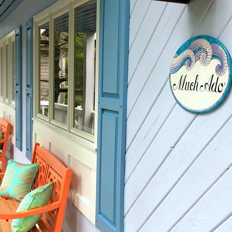 May include: A blue house with white trim and a blue shutter. There are two orange benches with cushions on the porch. A round sign with a mosaic design reads "Much Ado".