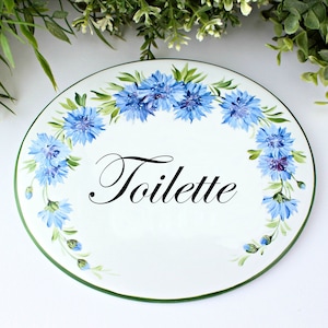 May include: White oval ceramic sign with a green border. The sign is decorated with blue cornflowers and the word "Toilette" in black cursive lettering.