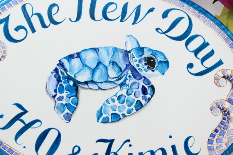 May include: A white oval sign with blue and gold mosaic trim. The sign says "The New Day" in blue cursive lettering. A blue painted sea turtle is in the center of the sign. Below the turtle, the sign says "Ho & Kimie" in blue cursive lettering.
