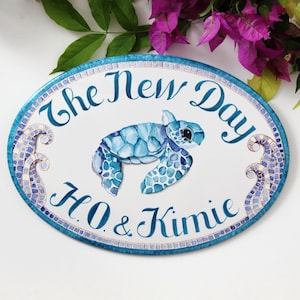 May include: Oval-shaped ceramic sign with a blue and white mosaic design. The sign features a painted sea turtle, waves, and the text "The New Day H.O. & Kimie".
