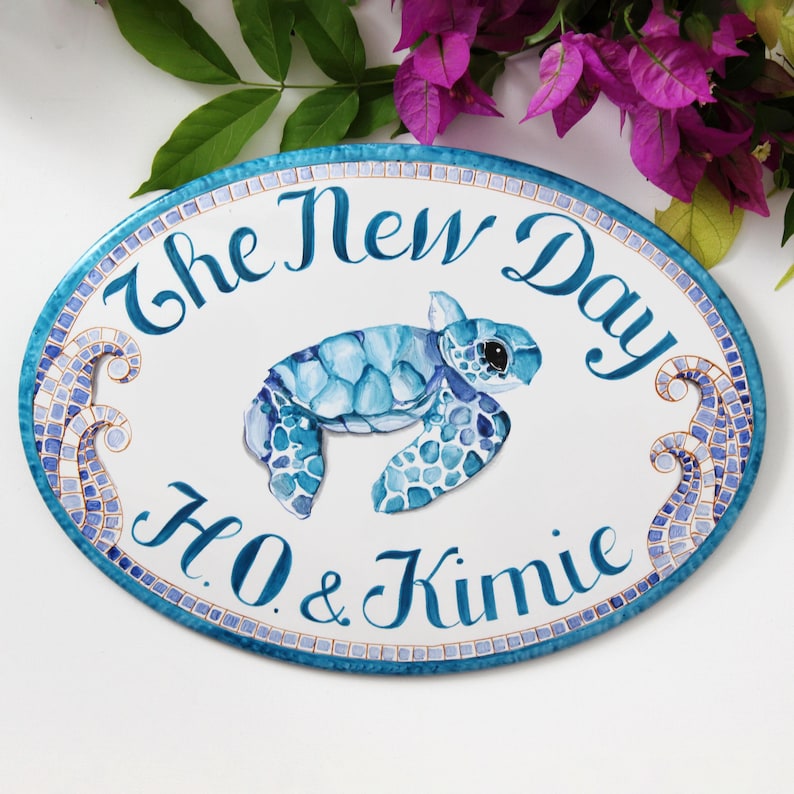 May include: Oval-shaped ceramic sign with a blue and white mosaic design. The sign features a painted sea turtle, waves, and the text "The New Day H.O. & Kimie".